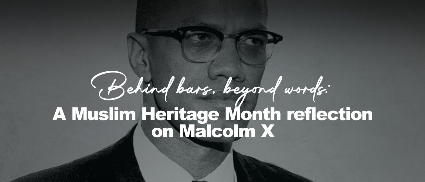 Black-and-white portrait of Malcolm X, shown from the chest up, wearing a dark suit, white shirt, and tie. He looks slightly to the right with a calm, serious expression, his round glasses catching soft light against a textured, light-colored background.